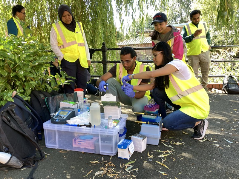 Hub colleagues crouch and stand around equipment from the 'Lab in a suitcase' whilst on the bank of the Ouseburn, preparing water samples for analysis